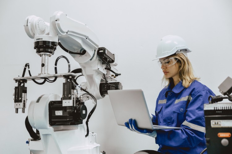 Female engineer in safety gear working on a laptop next to an industrial robot arm in a robotics lab