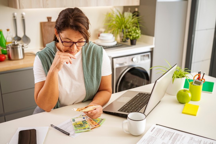 A woman managing her budget, carefully looking at dollar bills while working on her laptop in a modern kitchen