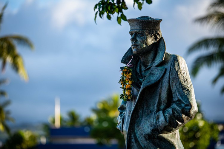 A bronze memorial sculpture of a sailor wearing a coat and hat, adorned with a flower garland, set against a tropical background