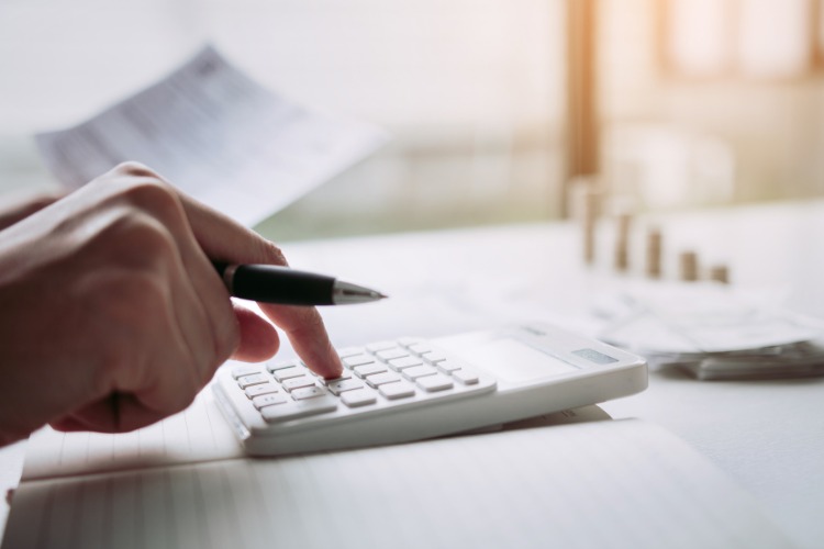 A person using a calculator while holding a pen, calculating financial expenses with documents in the background