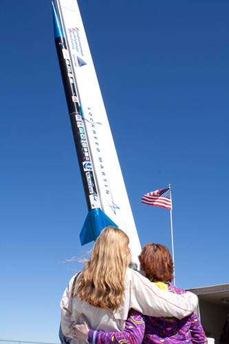 Two family members embracing while looking up at a Lockheed Martin memorial spaceflight rocket with an American flag in the background