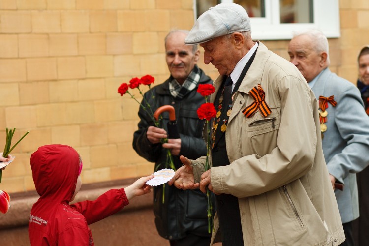 An elderly veteran receiving a handmade card from a young child during a Veterans Day event, surrounded by other veterans with flowers